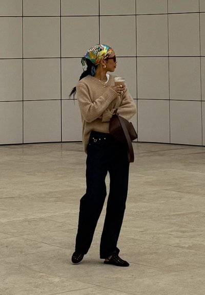 Suéter beige, jeans oscuros, pañuelo de cabeza estampado, sosteniendo una taza de café, de pie contra una pared de azulejos claros, con una bolsa marrón.