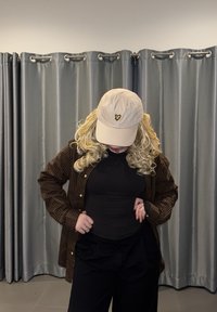 Casquette de baseball beige avec logo papillon, haut noir ajusté, chemise à carreaux marron, pantalon noir. En arrière-plan, des rideaux gris.