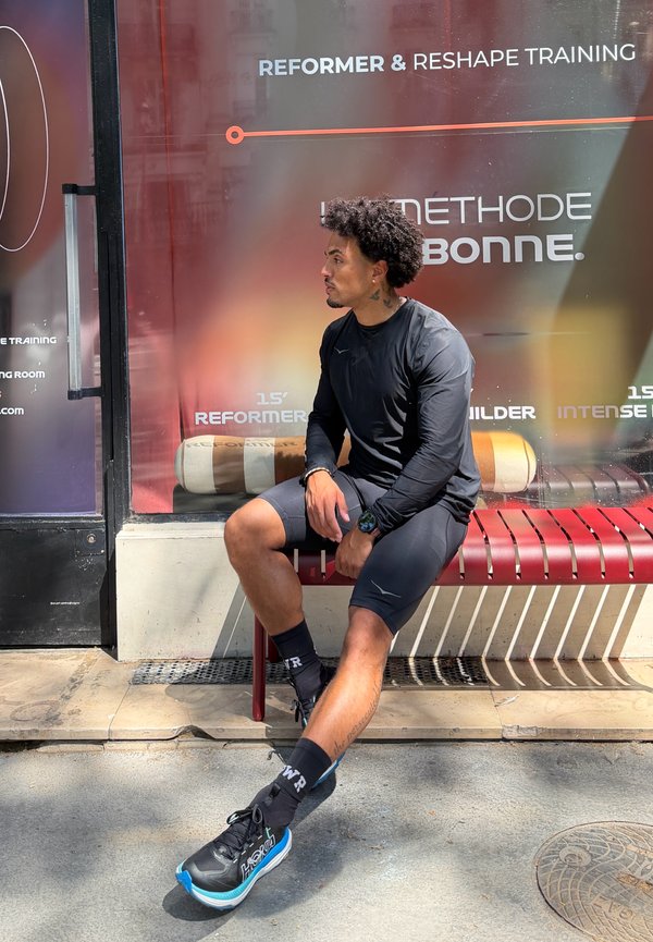Mand i sort sportsbeklædning, der sidder på en rød bænk udenfor et fitnessstudie med teksten "Reformer & Reshape Training" på vinduet bag ham.