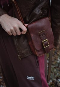 Bolso cruzado de cuero marrón texturizado con detalles en oro, asegurado con una hebilla. Llevado con una chaqueta con cremallera de color marrón oscuro y pantalones granate.