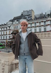 Jeune homme aux cheveux rose clair, portant un manteau brun à plusieurs couches et un sweat à capuche gris, debout sur des marches en ville, un bâtiment décoré et une rue visibles derrière lui.