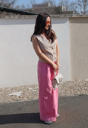 Mujer con chaleco beige sin mangas abotonado, pantalones anchos rosas, gafas de sol con tintes naranjas, sosteniendo un bolso blanco, de pie al aire libre sobre la acera.