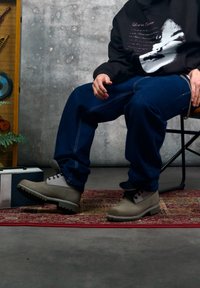 Bottines grises à cheville avec une tige texturée, des lacets noirs et une semelle robuste. Portées avec un pantalon en denim bleu et un sweat à capuche noir avec un motif graphique.