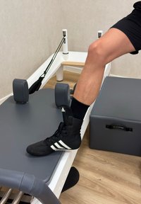 Sapatilha atlética preta com três riscas brancas, posicionada na borda de uma mesa de exercícios cinza acolchoada com bordas arredondadas.