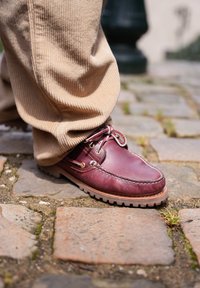 Καφέ παντελόνι από κορδούρα που καλύπτει τον αστράγαλο και ένα γυαλιστερό μπορντό δερμάτινο παπ ούτσι που στέκεται πάνω σε λιθόστρωτο.