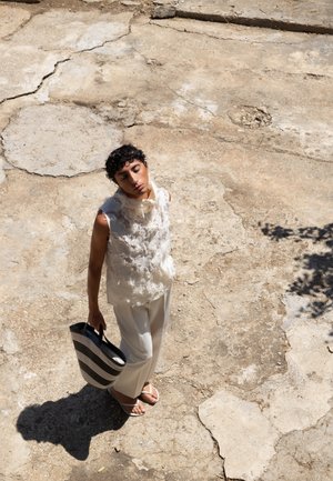 Top sin mangas, ligero y texturizado, con detalles en capas, combinado con pantalones blancos. Lleva un bolso de mano de rayas en blanco y negro. Fondo iluminado por el sol.