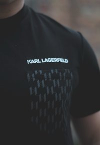 Melns kokvilnas T-krekls ar izteiktu balto "KARL LAGERFELD" logotipu un teksturētu melnu kabatu ar atkārtotu ģeometrisku rakstu.