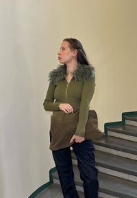 Zelený žebrovaný svetr s chlupatým límcem, kombinovaný s tmavými džínami. Drží hnědou strukturovanou kabelku. Neutrální pozadí se schody.