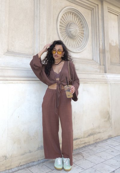 Top de punto suelto marrón con lazo al frente, combinado con pantalones de pierna ancha a juego. Lleva gafas de sol amarillas y zapatos verdes, sosteniendo una bebida.