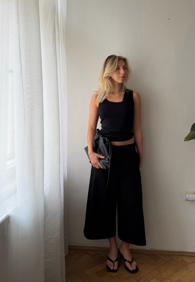 Mujer rubia de pie en interiores junto a una cortina blanca, lleva una camiseta de tirantes negra, pantalones negros anchos, sandalias negras y sostiene un bolso negro.