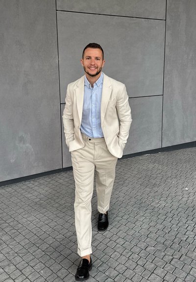 Traje beige claro con una chaqueta y pantalones a juego, combinado con una camisa azul claro y zapatos negros, con un fondo de pared y pavimento grises.