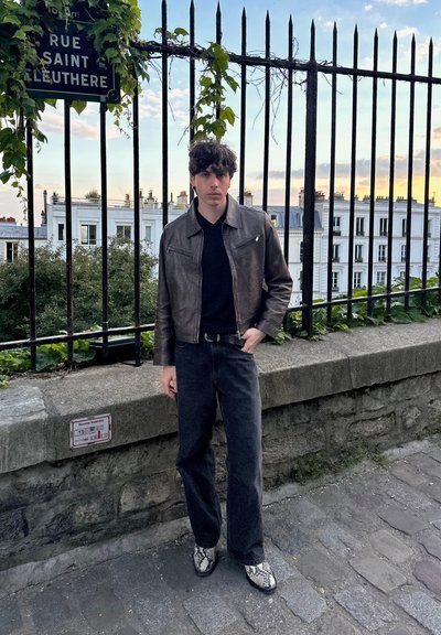 Chaqueta de cuero marrón, camisa negra de cuello en V, jeans oscuros de pierna ancha y botas con estampado de serpiente. De pie al lado de una pared de piedra y una cerca de hierro.