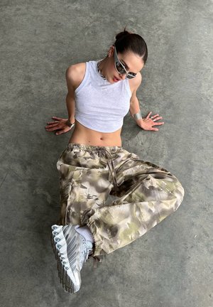 Fehér, rövidített pántos felső, kamuflázs mintájú cargo nadrág, ezüst sneaker cipők és elegáns napszemüveg. A modell a földön ült, kezét a megfelelő pozícióba helyezve.