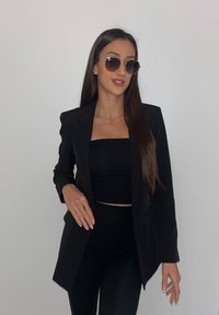 Blazer preto com design ajustado, tecido liso e lapelas afiadas, usado sobre um top curto preto e leggings. Acessorizado com óculos de sol escuros.