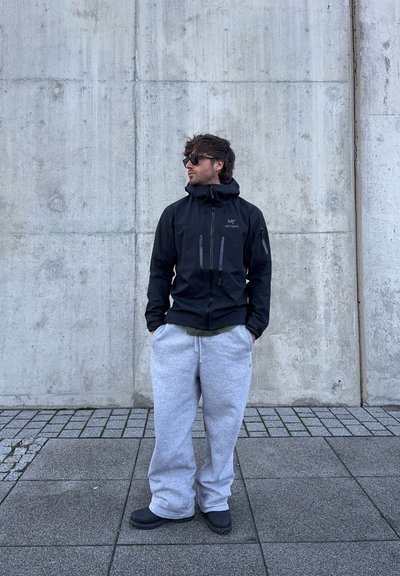 Chaqueta negra impermeable con capucha, bolsillos con cremallera y logotipo, combinada con pantalones deportivos gris claro y zapatillas de slip-on negras.