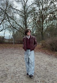 Giacca di pelle marrone sopra un maglione bordeaux, abbinata a jeans larghi azzurri. Sullo sfondo alberi spogli e un sentiero di ghiaia.