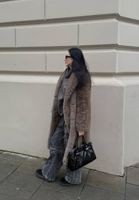 Ψεύτικο γούνινο παλτό σε καφέ χρώμα, γκρι φούτερ, χαλαρές γκρι τζιν παντελόνες και μαύρη τσάντα. Ά�τομο ντυμένο με μαύρα παπούτσια και γυαλιά ηλίου, περπατά δίπλα σε έναν τοίχο.