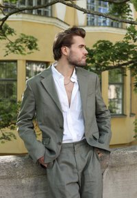 Traje de lino verde oliva claro con solapas de muesca, combinado con una camisa blanca desabrochada. De pie frente a una pared de concreto texturizada y vegetación.