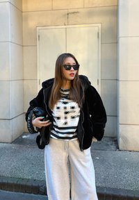 Chaqueta de pelaje sintético negro, camisa a rayas gris y blanca con estampado "92", pantalones de chándal grises, gafas de sol negras y un bolso de hombro.