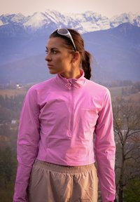 Jauna moteris rožinės striukės ir smėlio spalvos kelnių žvilgsnį kreipia į kairę, ant galvos dėvi saulės akinius, už jos matyti snieguotos kalnai ir miškinga kraštovaizdis.