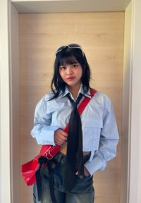 Camicia oversize azzurro chiaro con maniche lunghe, cravatta nera e borsa a tracolla rossa. Lo sfondo presenta pannelli in legno chiaro.