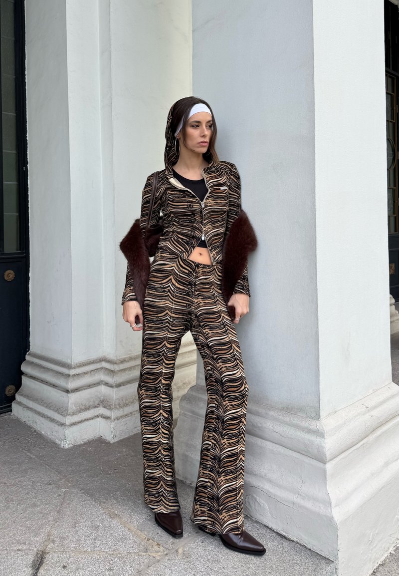 Frau trägt ein zweiteiliges Outfit mit Tigerstreifen und Fellärmeln, steht vor einem weißen Gebäude mit Säulen und schaut seitlich weg.