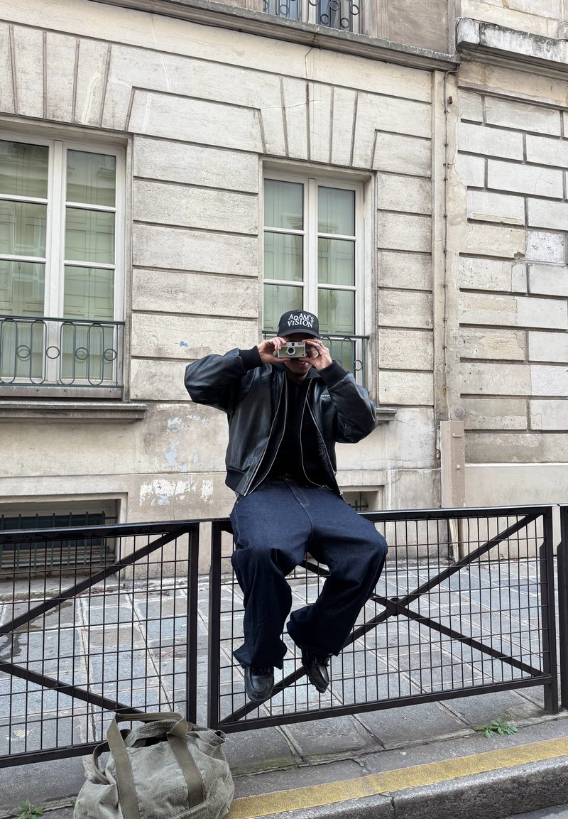 Person iført svart jakke og caps sitter på et metallgjerde, holder et kamera foran ansiktet, med en veske på fortauet i nærheten.