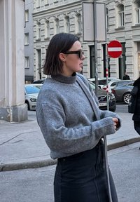 Šedý vlněný svetr s vysokým límcem, který má dlouhé, volné rukávy. V kombinaci s přiléhavou černou sukní je celý look doplněn tmavými slunečními brýlemi.