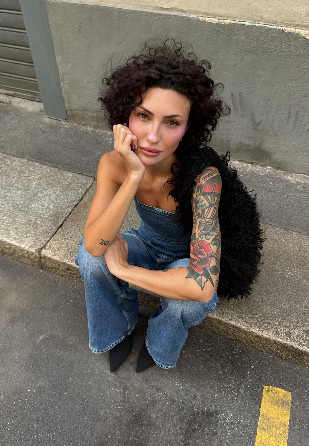 Kvinna med lockigt mörkt hår och färgglad ärm-tatuering sitter på en betongtrappa, klädd i jeans med vida ben och en svart texturerad topp.