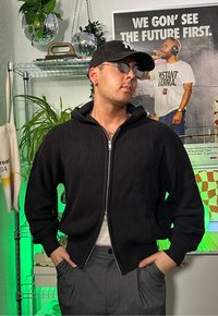 Schwarzer rippenstrukturierter Pullover mit Kapuze und vorderem Reißverschluss. Über einem hellen Hemd getragen, ergänzt durch graue Hose und Sonnenbrille.