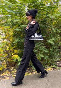 Top negro de manga larga, pantalones anchos negros, gorro negro, botines negros de tacón alto y un bolso transparente con detalles blancos. Vegetación verde de fondo.