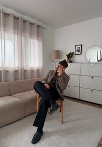 Άνδρας με γυαλιά και καπέλο κάθεται σε ξύλινη καρέκλα σε μοντέρνο σαλόνι με μπεζ καναπέ, επιδαπέδιο φωτιστικό, ντουλάπι, φυτό και στρογγυλό καθρέφτη τοίχου.