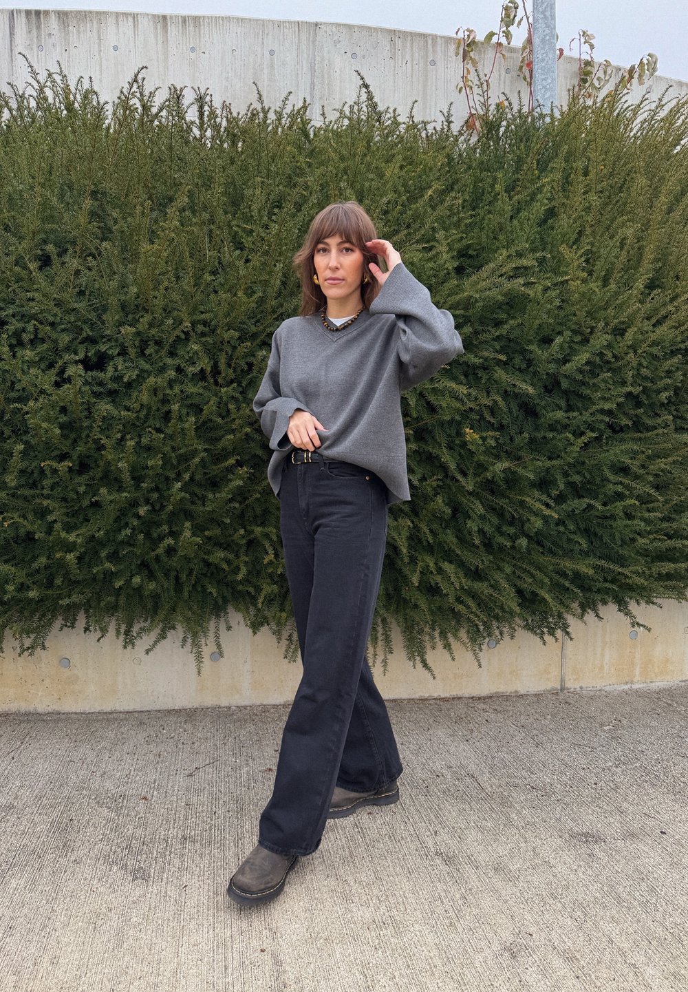 Maglione grigio oversize con maniche larghe, abbinato a pantaloni neri a vita alta e svasati, e stivali scuri. Sullo sfondo un cespuglio verde.