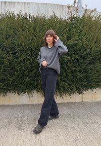 Maglione grigio oversize con maniche larghe, abbinato a pantaloni neri a vita alta e svasati, e stivali scuri. Sullo sfondo un cespuglio verde.