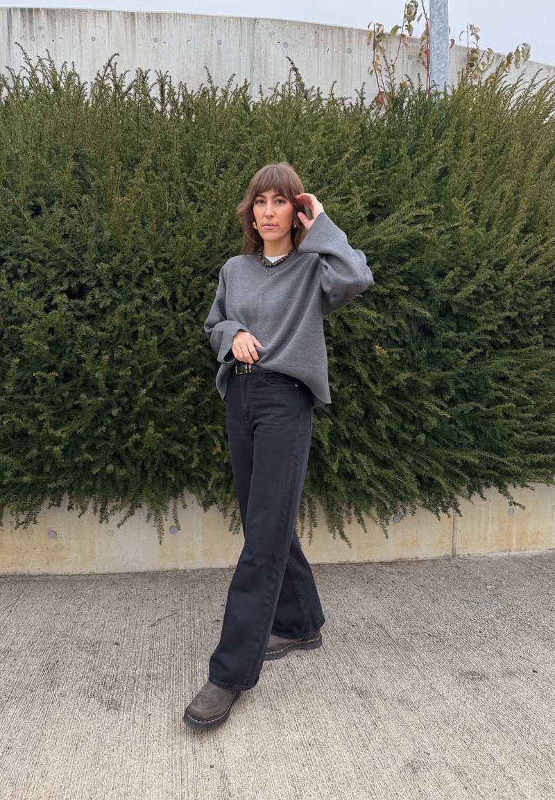 Grå oversized genser med vide ermer, kombinert med høytlivede svarte vide bukser og mørke støvler. Mot en bakgrunn av grønne busker.