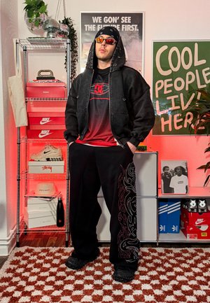Joven con sudadera negra con capucha, camiseta roja de DC, pantalones negros con diseño de llamas, gafas de sol, de pie en una habitación con zapatillas de marca y carteles motivacionales.