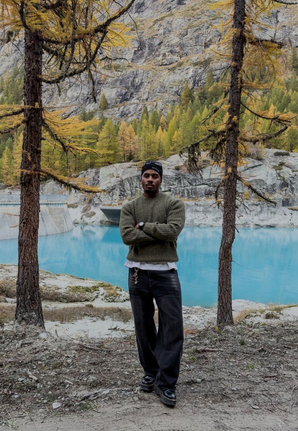 Un homme se tient les bras croisés au bord d’un lac de montagne turquoise, encadré par des arbres aux couleurs automnales et des falaises rocheuses.