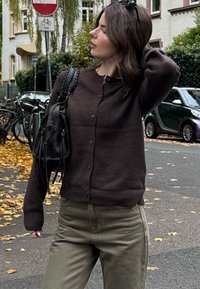 Ruskea neulottu neuletakki napillaisella suljennalla, yhdistetty oliivinvihreisiin housuihin. Malli pitää kädessään pientä mustaa käsilaukkua. Kaupallinen syysympäristö pudonneiden lehtien keskellä.