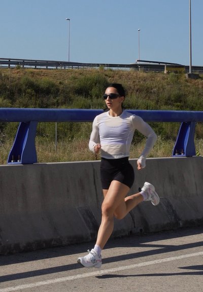 Mujer con gafas de sol y ropa deportiva corriendo por una carretera junto a una barrera de hormigón, con una ladera cubierta de hierba y una autopista al fondo.