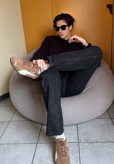 Zapatillas de ante marrón, vaqueros negros y una camisa oscura, sentado en un puff ligero de color gris con una textura suave. La pared es de madera clara.