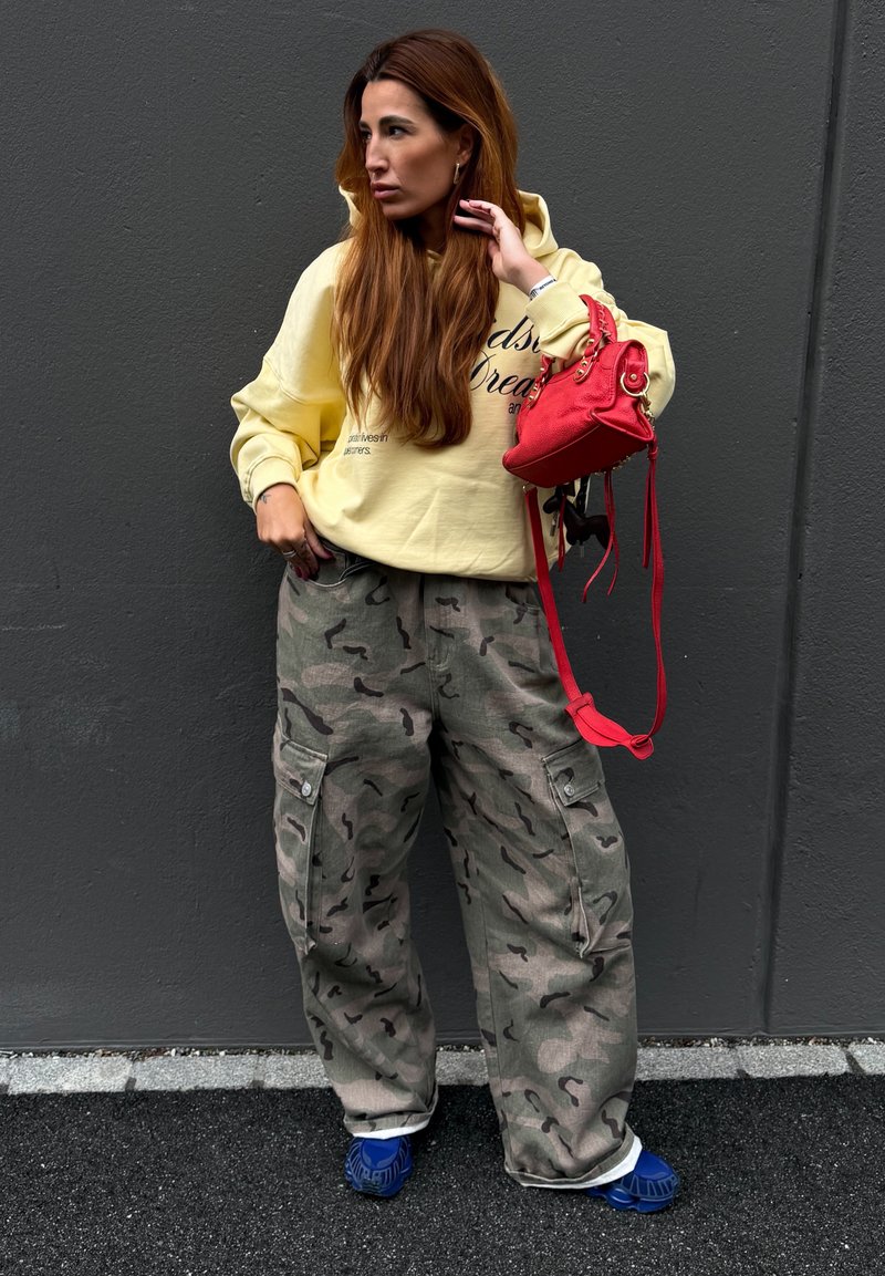 Kvinde med langt brunt hår iført gul hættetrøje, camouflage cargo bukser, blå sko, og holder en rød håndtaske mod en mørk væg.