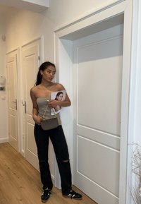 Lichtgrijze fitted strapless top, zwarte ripped jeans, zwarte schoenen met gouden accenten, kleine bruine schoudertas, een tijdschrift vasthoudend, eenvoudige witte deur.