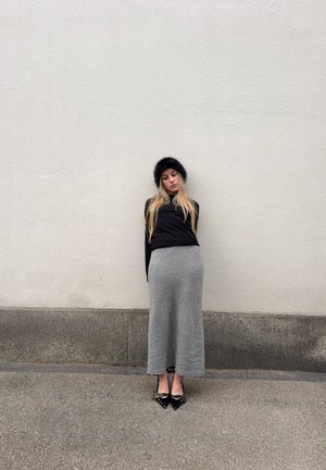 Joven con cabello largo y rubio, gorro de piel negro, blusa negra, falda larga gris y zapatos negros puntiagudos, de pie contra una pared lisa y clara.