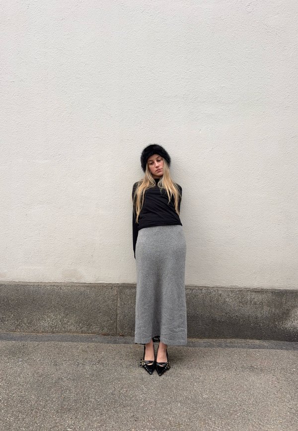 Νεαρή γυναίκα με μακριά ξανθά μαλλιά, μαύρο γούνινο καπέλο, μαύρη μπλούζα, γκρι μακριά φούστα και μαύρα μυτερά παπούτσια στέκεται μπροστά σε ένα απλό ανοιχτό τοίχο.