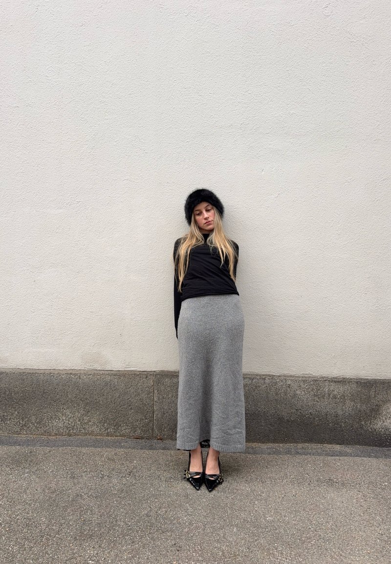 Νεαρή γυναίκα με μακριά ξανθά μαλλιά, μαύρο γούνινο καπέλο, μαύρη μπλούζα, γκρι μακριά φούστα και μαύρα μυτερά παπούτσια στέκεται μπροστά σε ένα απλό ανοιχτό τοίχο.