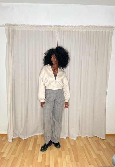 Chaqueta corta de cremallera blanca sobre un pantalón gris de cuadros de gran tamaño. Zapatos de plataforma negros. El fondo presenta cortinas de un beige claro.