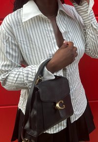 Borsa a tracolla nera in pelle con chiusura in oro. Modella indossa una camicia bianca a righe con colletto ampio e una gonna nera. Sfondo rosso.