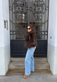 Hnedá krátka bunda z umelej kožušiny, svetlomodré široké džínsové nohavice a kožené topánky s platformou v hnedej farbe, na pozadí dekoratívna čierna kovaná brána.