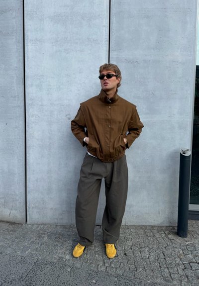 Hombre con chaqueta marrón, pantalones anchos grises, zapatos amarillos y gafas de sol negras, de pie con las manos en los bolsillos frente a una pared gris.