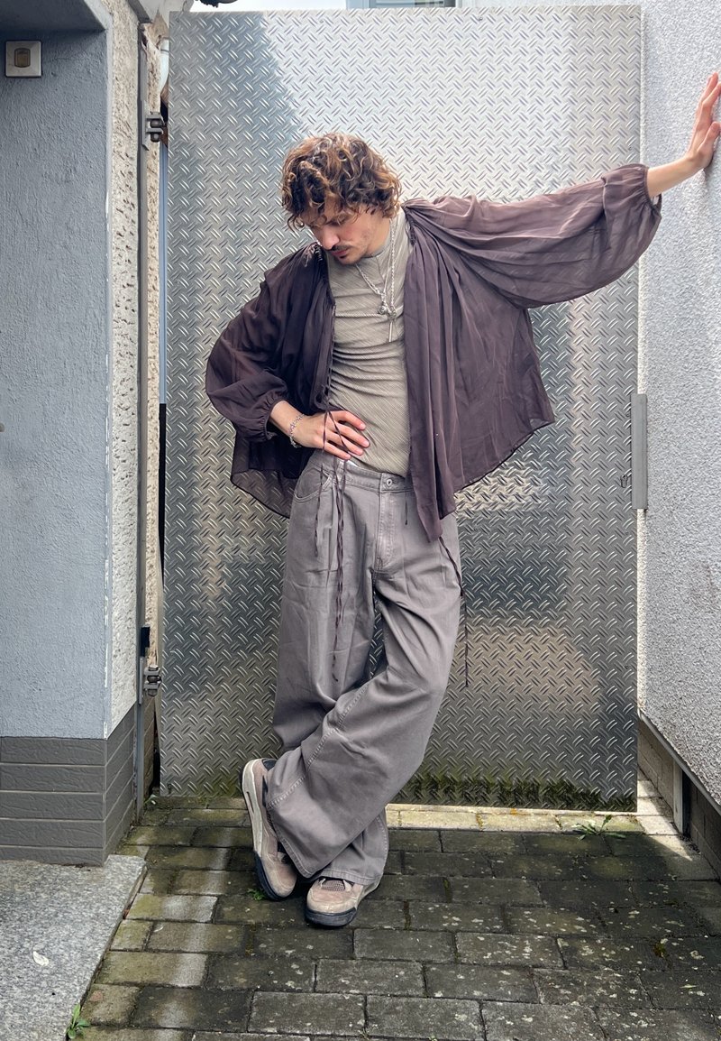 Homme aux cheveux bouclés portant un pantalon gris ample, un haut beige et une veste sombre transparente, appuyé contre un mur métallique dans une ruelle extérieure étroite.